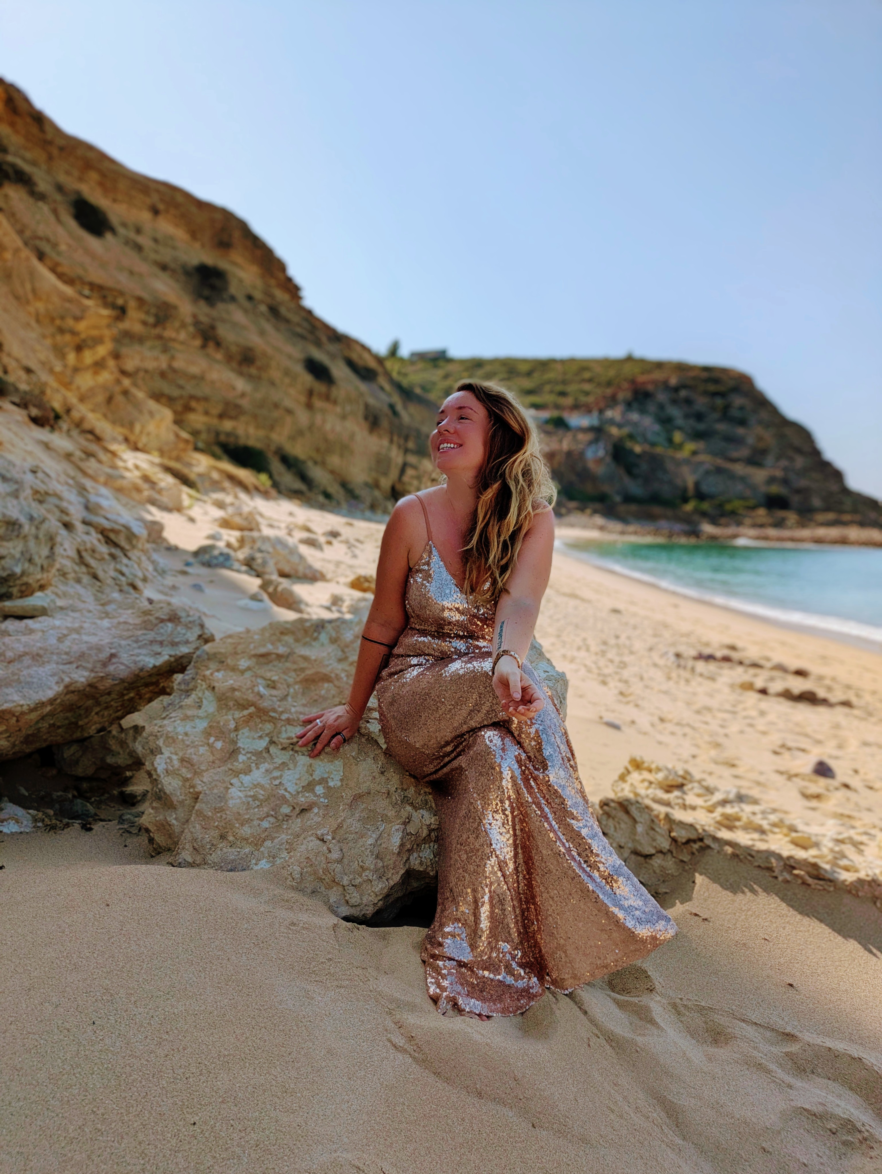Women in golden ball gown sits on a rock on the beach